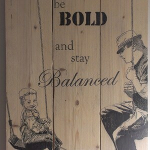 May include: A wooden sign with the text "be BOLD and stay Balanced" printed in black. The sign features a black and white illustration of a child on a swing and a man standing next to the swing.