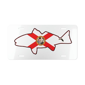 May include: White license plate with a black outline of a redfish. The redfish is filled with the Florida state flag, which features a red cross on a white background with the state seal in the center.