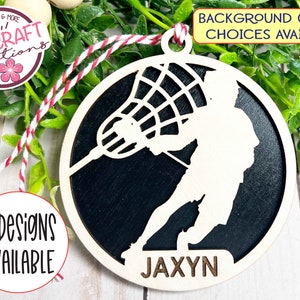 May include: A white wooden lacrosse player ornament with a black background. The ornament is round and has a hole at the top for hanging. The player is holding a lacrosse stick and the ball is in the net. The name "JAXYN" is written at the bottom of the ornament.