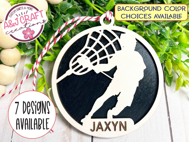 May include: A white wooden lacrosse player ornament with a black background. The ornament is round and has a hole at the top for hanging. The player is holding a lacrosse stick and the ball is in the net. The name "JAXYN" is written at the bottom of the ornament.