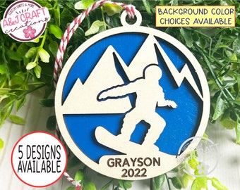 May include: A wooden Christmas ornament featuring a snowboarder silhouette against a blue mountain scene. The ornament has a natural wood finish with a blue background and the name "Grayson" and the year "2022" engraved. It includes a red and white string for hanging.