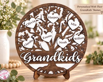 May include: A circular wooden display featuring a dark brown laser-cut tree silhouette against a white background. Six white heart-shaped cutouts are attached to the branches, each inscribed with a name. The word Grandkids is written in white script at the base of the tree trunk. The piece is supported by a small wooden stand.
