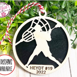 May include: A wooden lacrosse player ornament with a black background. The ornament is round and has a string for hanging. The player is holding a lacrosse stick and the net is visible. The text "HEYDT #19 2022" is printed on the ornament.