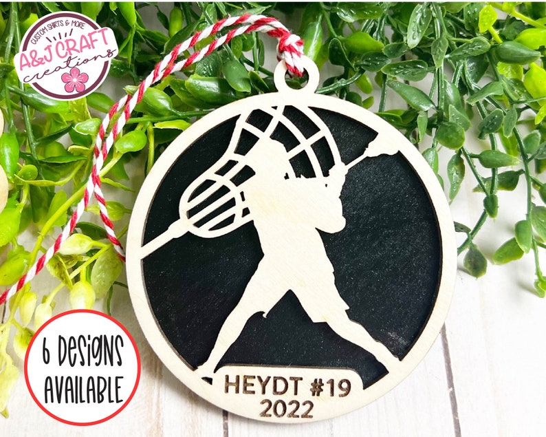 May include: A wooden lacrosse player ornament with a black background. The ornament is round and has a string for hanging. The player is holding a lacrosse stick and the net is visible. The text "HEYDT #19 2022" is printed on the ornament.