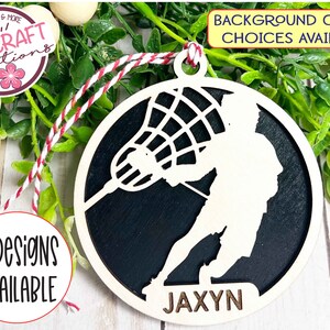 May include: A wooden lacrosse player ornament with a black background and the name "JAXYN" written on the bottom. The ornament is round and has a string attached to the top for hanging.