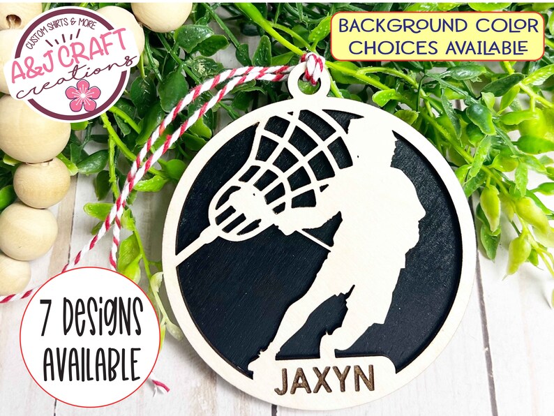 May include: A wooden lacrosse player ornament with a black background and the name "JAXYN" written on the bottom. The ornament is round and has a string attached to the top for hanging.