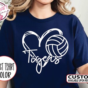 May include: Navy blue t-shirt featuring a white heart-shaped design with a volleyball and the word "Tigers". The shirt also has the text "SELECT TSHIRT COLOR" and "CUSTOM AVAILABLE UPON REQUEST".
