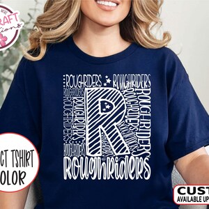 May include: Navy blue t-shirt with a white graphic design. The design features the word "ROUGHRIDERS" in various orientations, surrounding a large letter "R" with a striped pattern and stars. The shirt also has the text "SELECT TSHIRT COLOR" and "CUSTOM AVAILABLE UPON REQUEST".