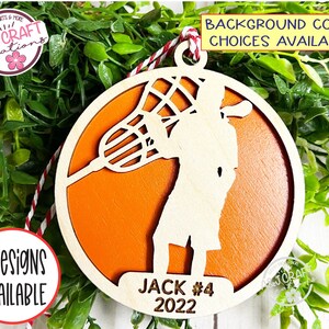 May include: A wooden orange lacrosse player ornament with a white silhouette of a player holding a lacrosse stick. The ornament has the text "JACK #4 2022" on the bottom.
