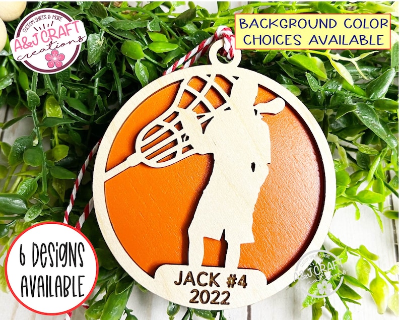 May include: A wooden orange lacrosse player ornament with a white silhouette of a player holding a lacrosse stick. The ornament has the text "JACK #4 2022" on the bottom.