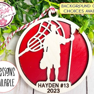 May include: A red and white wooden lacrosse player ornament with the name Hayden #13 and the year 2023. The ornament is a silhouette of a lacrosse player holding a lacrosse stick and net.