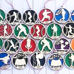 May include: A collection of 18 wooden ornaments featuring various sports and activities. Each ornament is painted in different colors and includes a name and year. The ornaments are perfect for decorating a Christmas tree or adding a personal touch to any space.