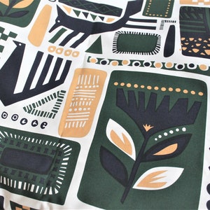 Op de afbeelding: Een witte stof met een herhalend patroon van zwarte, groene en gouden geometrische vormen. Het patroon omvat gestileerde bloemen, vogels en abstracte ontwerpen. De stof heeft de tekst "MARIMEKKO OYJ SUOMI-FINLAND SANNA ANNUKKA 2017: "SVAALE" 100% CO WA品" gedrukt aan de rechterkant.