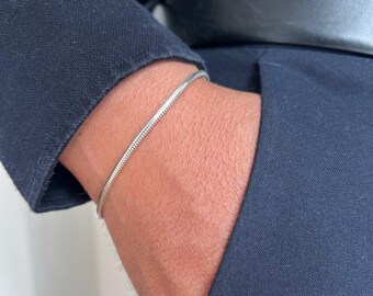 May include: A silver snake chain bracelet is worn on a wrist, with a dark blue jacket and pants in the background. The bracelet is a simple, elegant design, perfect for everyday wear. The bracelet is approximately 7 inches in length.