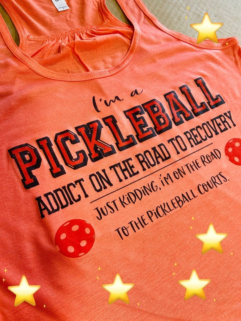 May include: Orange tank top with the text "I'm a Pickleball Addict on the Road to Recovery. Just Kidding, I'm on the Road to the Pickleball Courts."  The text is in black with a red outline. There are two red pickleball paddles with white dots on the shirt.
