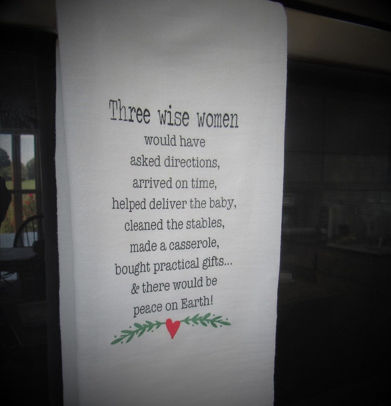 May include: A white kitchen towel with a black and white text print that reads "Three wise women would have asked directions, arrived on time, helped deliver the baby, cleaned the stables, made a casserole, bought practical gifts... & there would be peace on Earth!" with a red heart and green leaves.
