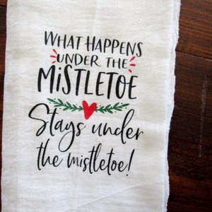May include: White kitchen towel with black text that reads "What happens under the mistletoe stays under the mistletoe!" with a red heart and green leaves.