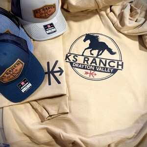 May include: A tan hoodie with a black silhouette of a horse and the text "KS Ranch Drayton Valley" in a circle. Three trucker hats with black mesh and a brown leather patch with the text "KS Ranch Drayton Valley" are also in the image.