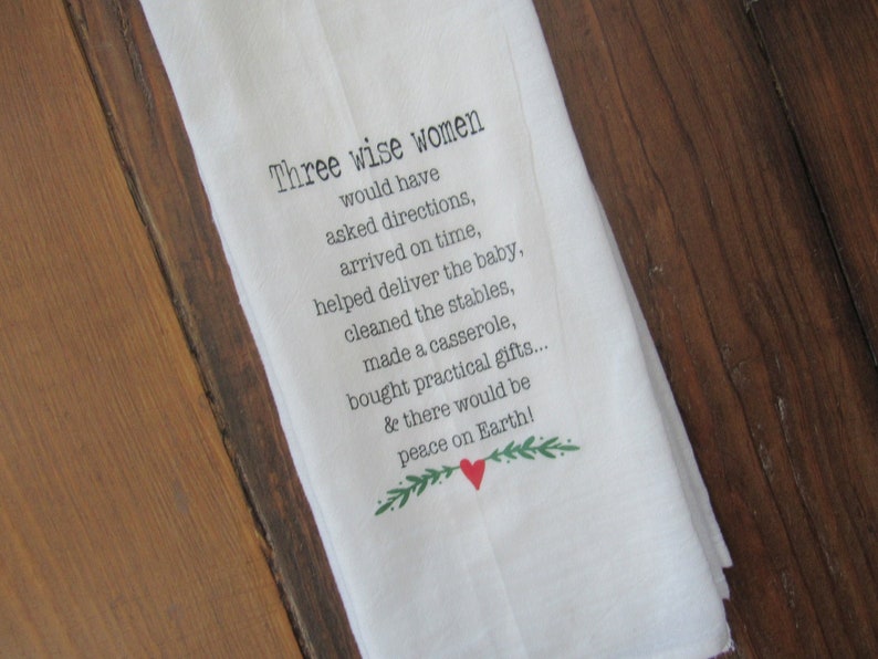 May include: White kitchen towel with a humorous Christmas-themed saying: "Three wise women would have asked directions, arrived on time, helped deliver the baby, cleaned the stables, made a casserole, bought practical gifts... & there would be peace on Earth!" The text is in black with a green sprig and a red heart.