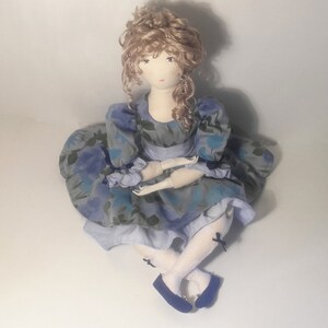 May include: A handmade fabric doll with blonde curly hair, wearing a blue and white floral dress with a white lace trim and black bows on her shoes.