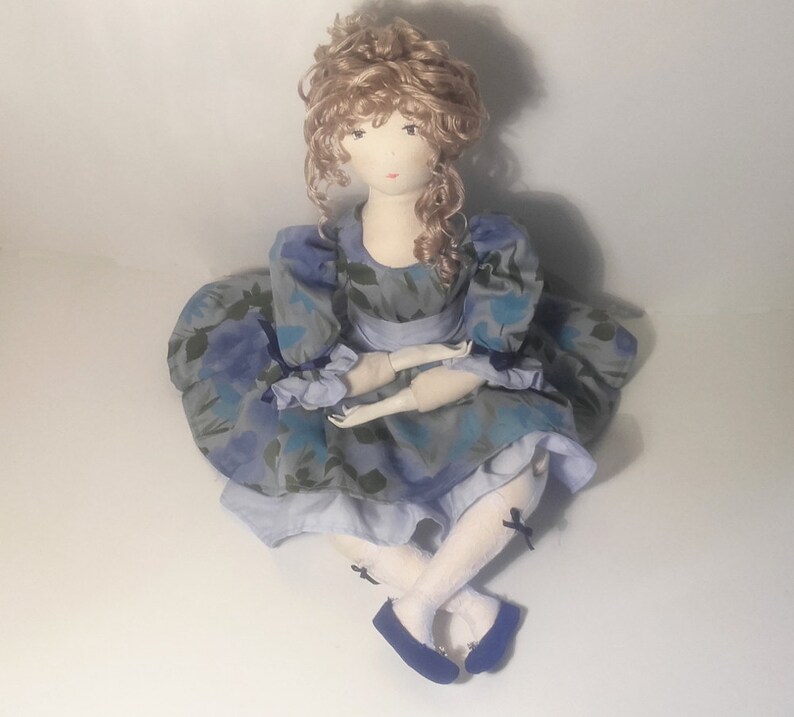 May include: A handmade fabric doll with blonde curly hair, wearing a blue and white floral dress with a white lace trim and black bows on her shoes.