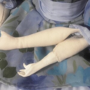 May include: A close-up of a doll's arms, showing the fabric construction of the arms and hands. The doll is wearing a blue dress with a floral pattern.