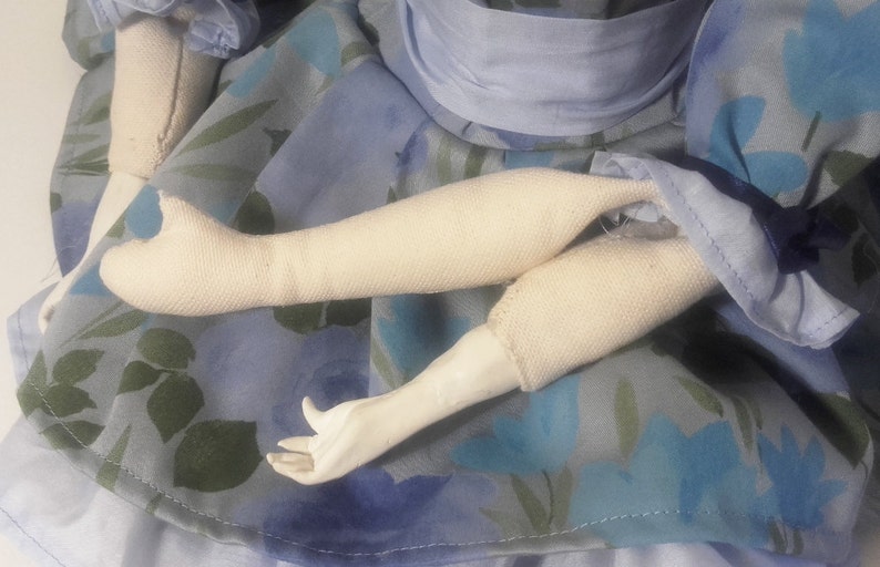May include: A close-up of a doll's arms, showing the fabric construction of the arms and hands. The doll is wearing a blue dress with a floral pattern.