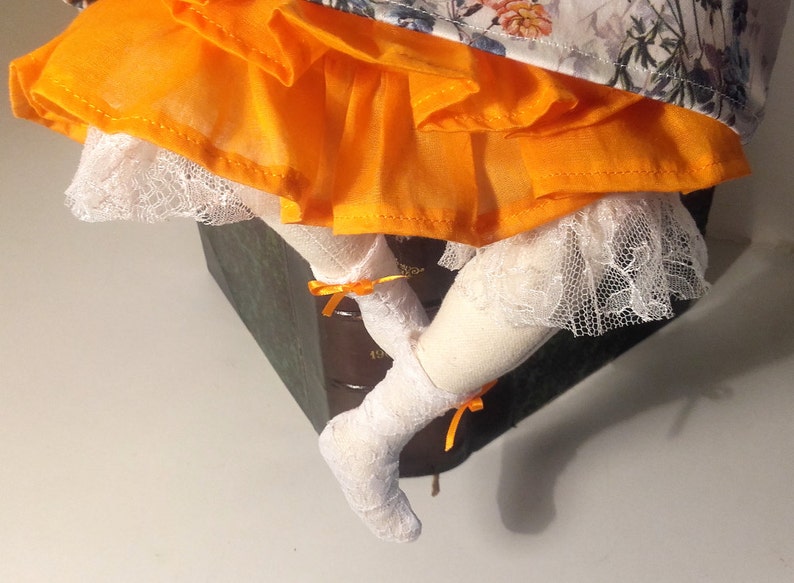 May include: A doll with white lace socks and orange ruffled skirt sitting on a stack of books.