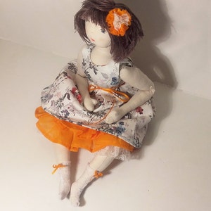 May include: A handmade doll with brown hair and a white dress with orange and floral accents. The doll is wearing a small orange flower on her hair.