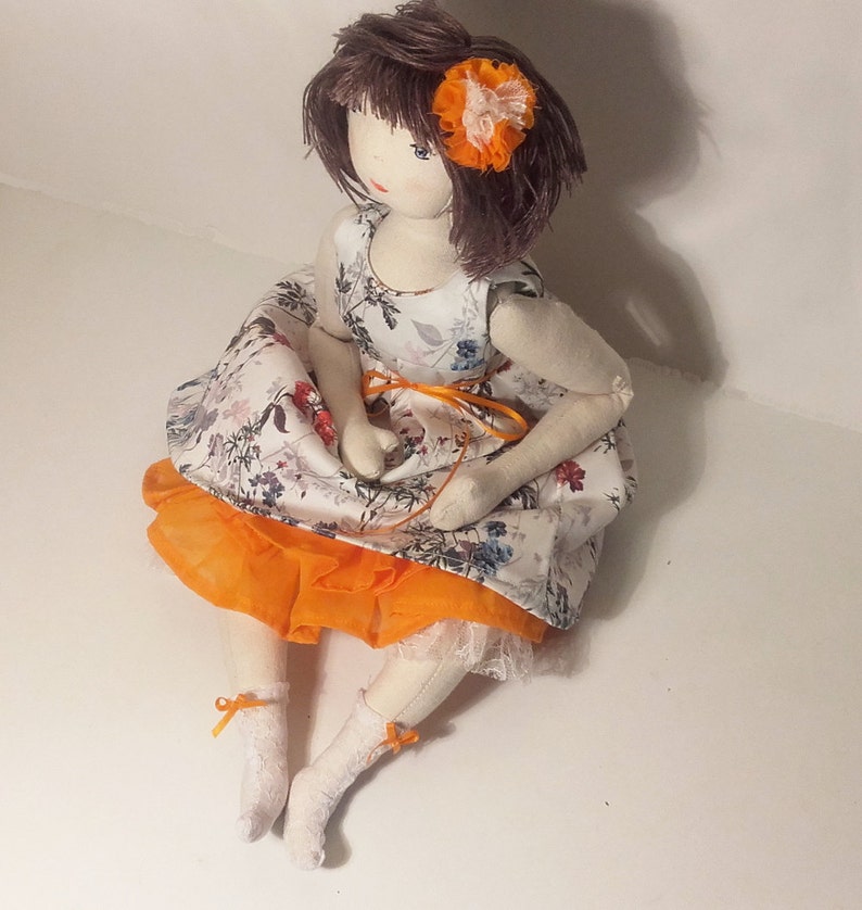 May include: A handmade doll with brown hair and a white dress with orange and floral accents. The doll is wearing a small orange flower on her hair.