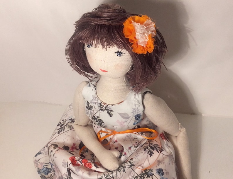 May include: A handmade doll with brown hair, wearing a white dress with a floral pattern and an orange flower in her hair.