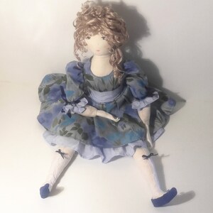 May include: A handmade doll with curly blonde hair wearing a blue and green floral dress with a blue sash and white socks and blue shoes.