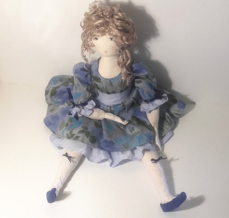 May include: A handmade doll with curly blonde hair wearing a blue and green floral dress with a blue sash and white socks and blue shoes.