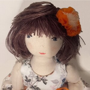 May include: A handmade cloth doll with brown yarn hair, blue eyes, and a red painted mouth. The doll is wearing a white dress with a floral pattern and an orange flower in her hair.