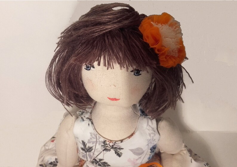 May include: A handmade cloth doll with brown yarn hair, blue eyes, and a red painted mouth. The doll is wearing a white dress with a floral pattern and an orange flower in her hair.