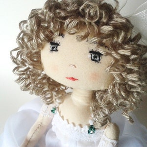 May include: A handmade doll with curly blonde hair, wearing a white dress with lace trim and green embellishments. The doll has embroidered eyes and a red mouth.