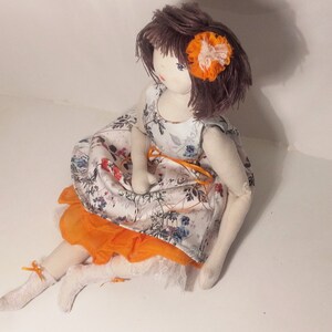 May include: A handmade fabric doll with brown hair, wearing a white dress with floral print and an orange skirt. The doll has a flower in its hair.