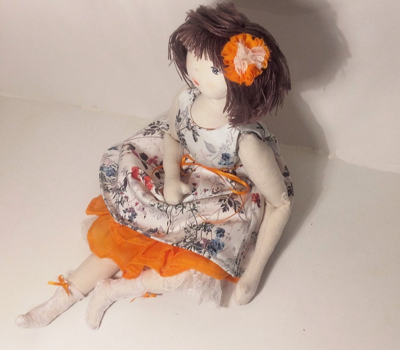 May include: A handmade fabric doll with brown hair, wearing a white dress with floral print and an orange skirt. The doll has a flower in its hair.