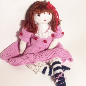 May include: A knitted doll with brown hair, wearing a pink dress with red flower accents, white lace collar, white socks, and black and white striped stockings.