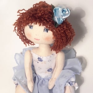 May include: A handmade doll with curly red hair, blue eyes, and a light blue dress with a floral pattern. The doll is wearing a light blue flower on her head.