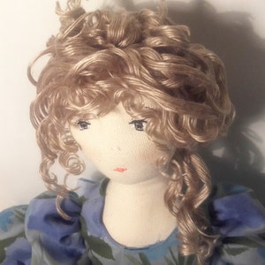 May include: A handmade doll with a white fabric body and a blonde curly wig. The doll is wearing a blue and green floral dress.