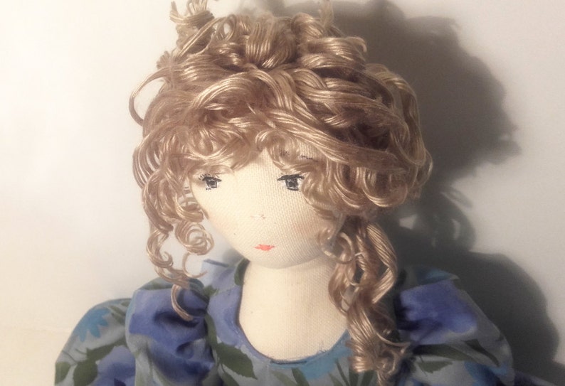 May include: A handmade doll with a white fabric body and a blonde curly wig. The doll is wearing a blue and green floral dress.