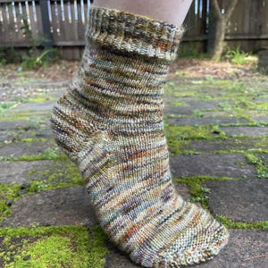 May include: A hand-knitted ankle sock featuring a variegated yarn pattern with earthy tones of olive green, brown, tan, and subtle hints of orange and blue. The sock has a ribbed cuff at the top and a consistent stockinette stitch throughout.