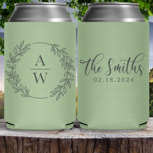 May include: Two green can coolers with a wedding theme. The cooler on the left has a wreath with leaves and the initials 'A W' inside. The cooler on the right has the text 'The Smiths' and the date '02.18.2024'.