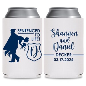 May include: Two white can coolers with blue text and graphics. The left cooler features a silhouette of a police officer carrying a bride over his shoulder. The text reads 'Sentenced to Life!' with a blue shield with the letter 'D' inside. The right cooler features the text 'Shannon and Daniel Decker 03.17.2024'.
