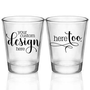 May include: Two clear glass shot glasses. One shot glass has the text 'your custom design here' in black cursive font. The other shot glass has the text 'here too' in black cursive font.