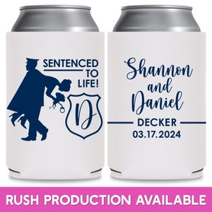 May include: Two white can coolers with blue text and graphics. The left cooler has a silhouette of a police officer carrying a woman over his shoulder. The text reads 'Sentenced to Life!' and 'D'. The right cooler has the text 'Shannon and Daniel Decker 03.17.2024'. Both coolers are for wedding favors.