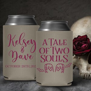 May include: Two tan can coolers with pink text. The cooler on the left says 'Kelsey & Dave OCTOBER 28TH, 2022'. The cooler on the right says 'A TALE OF TWO SOULS' with two tombstones with hearts and bats on either side.
