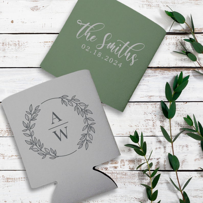 May include: Two personalized can coolers. One cooler is gray with a wreath design and the initials 'A/W' in the center. The other cooler is green with the text 'the Smiths 02.18.2024'.