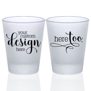 May include: Two frosted shot glasses. The shot glass on the left has the text 'your custom design here' in black cursive font. The shot glass on the right has the text 'here too' in black cursive font.